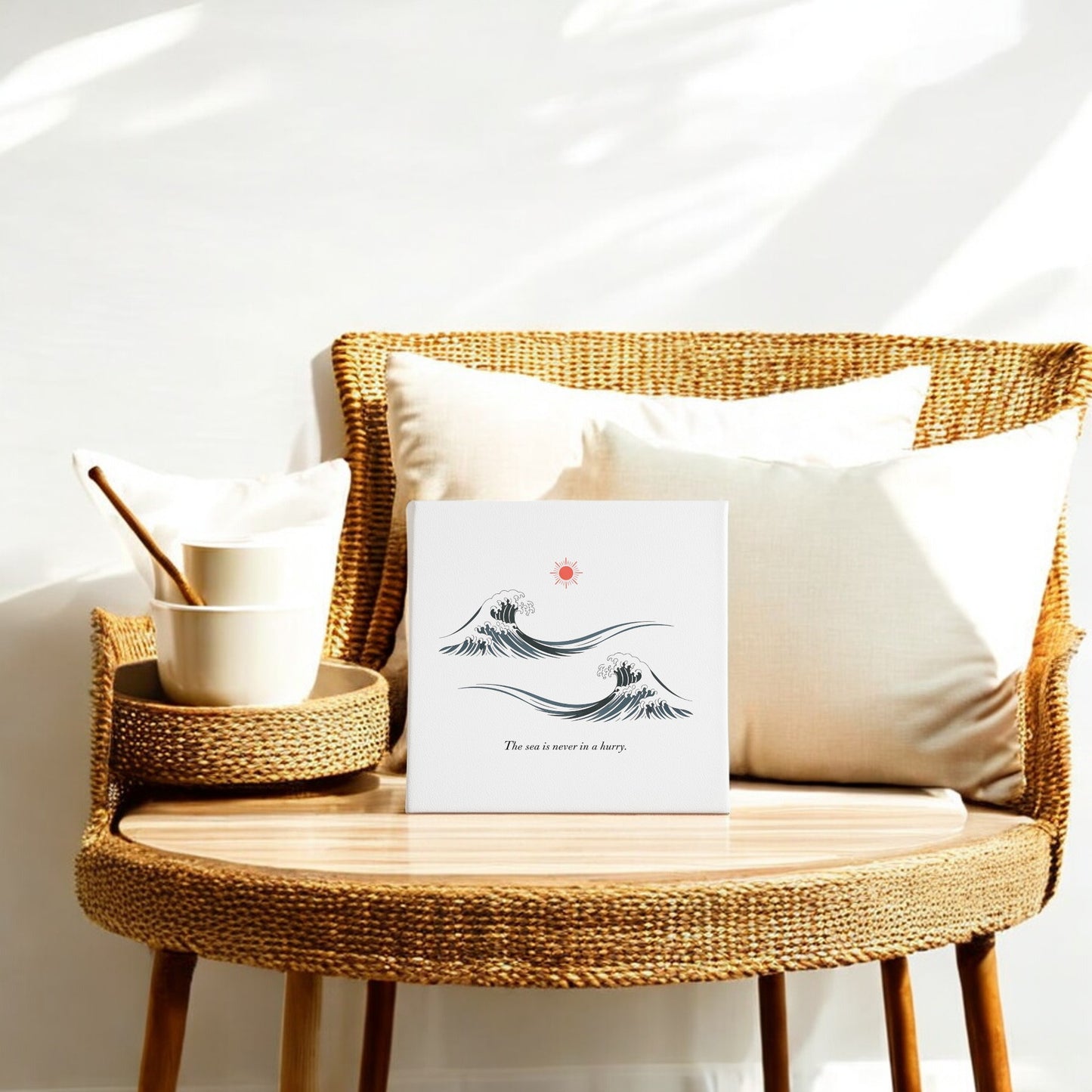A rattan chair with white cushions sits beside a wooden table topped with stacked bowls and Mirabilia Boutique’s "The Sea Is Never in a Hurry Canvas – Serene Ocean Calm." Sunlight casts soft shadows on the wall behind.