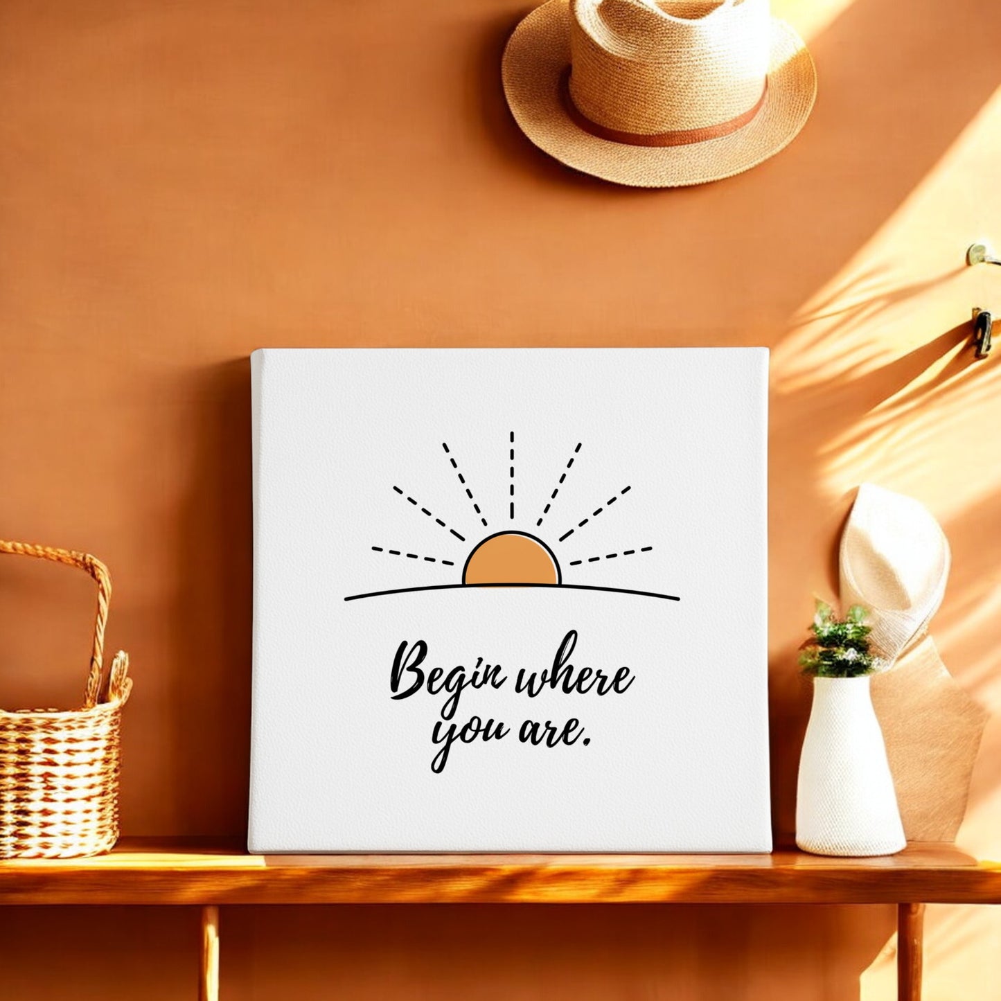 The Mirabilia Boutique "Begin Where You Are Canvas – Sunrise Wall Affirmation" features a simple sun illustration and motivational phrase, displayed on a wooden shelf with woven baskets, a plant, and straw hats against a warm brown wall.