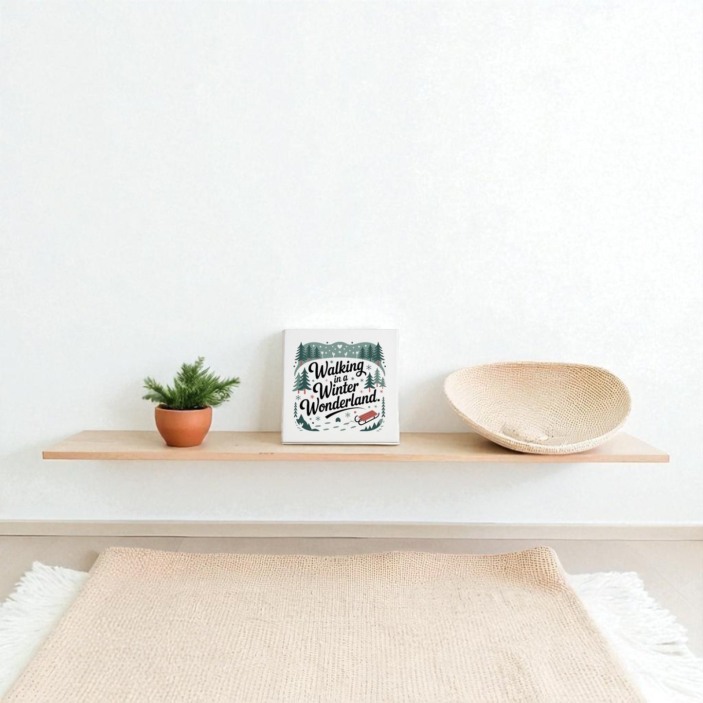 A minimalist floating shelf showcases a small potted plant, a woven basket, and the Mirabilia Boutique Winter Wonderland Canvas | 8x8 Holiday Decor against a plain white wall above a beige rug.