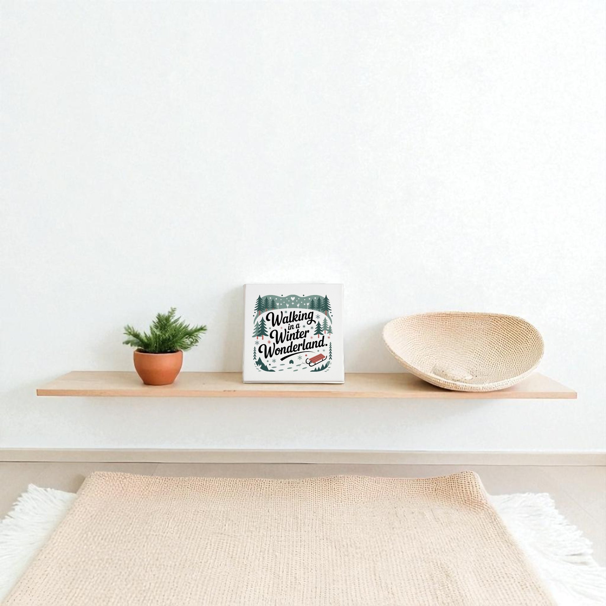A minimalist floating shelf showcases a small potted plant, a woven basket, and the Mirabilia Boutique Winter Wonderland Canvas | 8x8 Holiday Decor against a plain white wall above a beige rug.