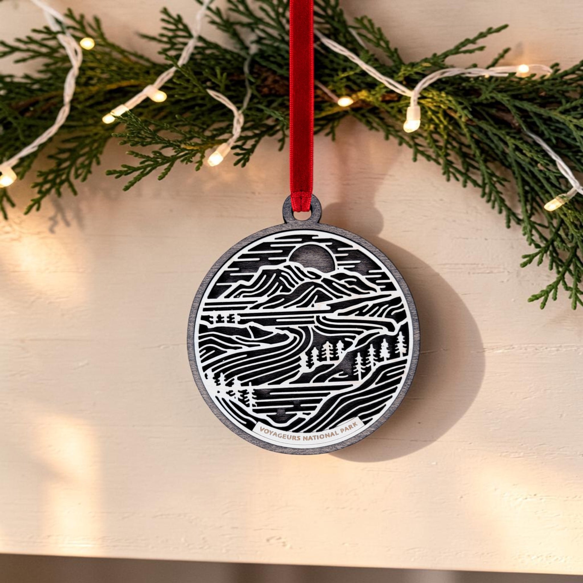 The Mirabilia Boutique Voyageurs National Park Line Art Christmas Ornament is an oval, handcrafted wooden piece featuring layered wood art with a black and white scenic design of trees and water, plus "Voyageurs National Park" text at the bottom. It hangs from a red ribbon, while pine branches with string lights are shown above.