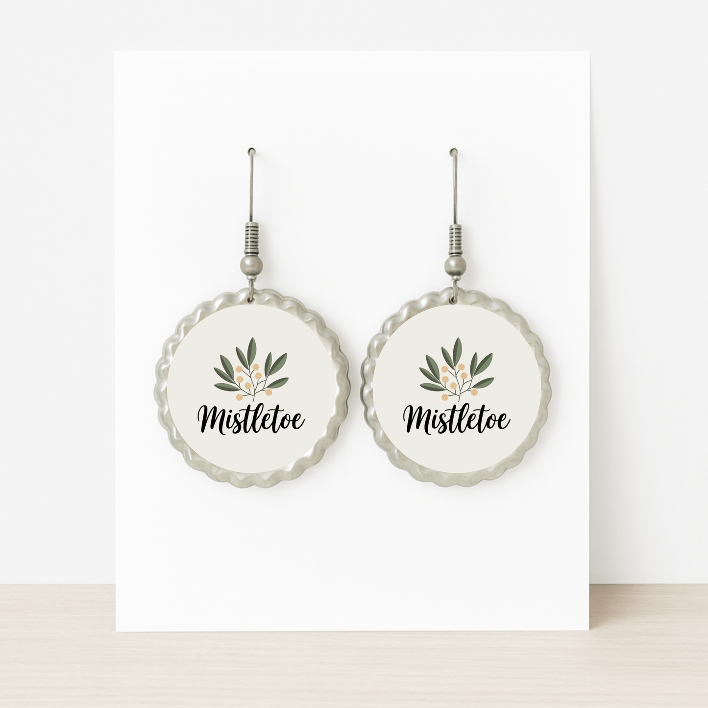 Mirabilia Boutique's Mistletoe Holiday Earrings are round, hypoallergenic studs with scalloped silver edges, featuring a mistletoe illustration and "Mistletoe" in cursive—festive botanical Christmas jewelry displayed on a white card.