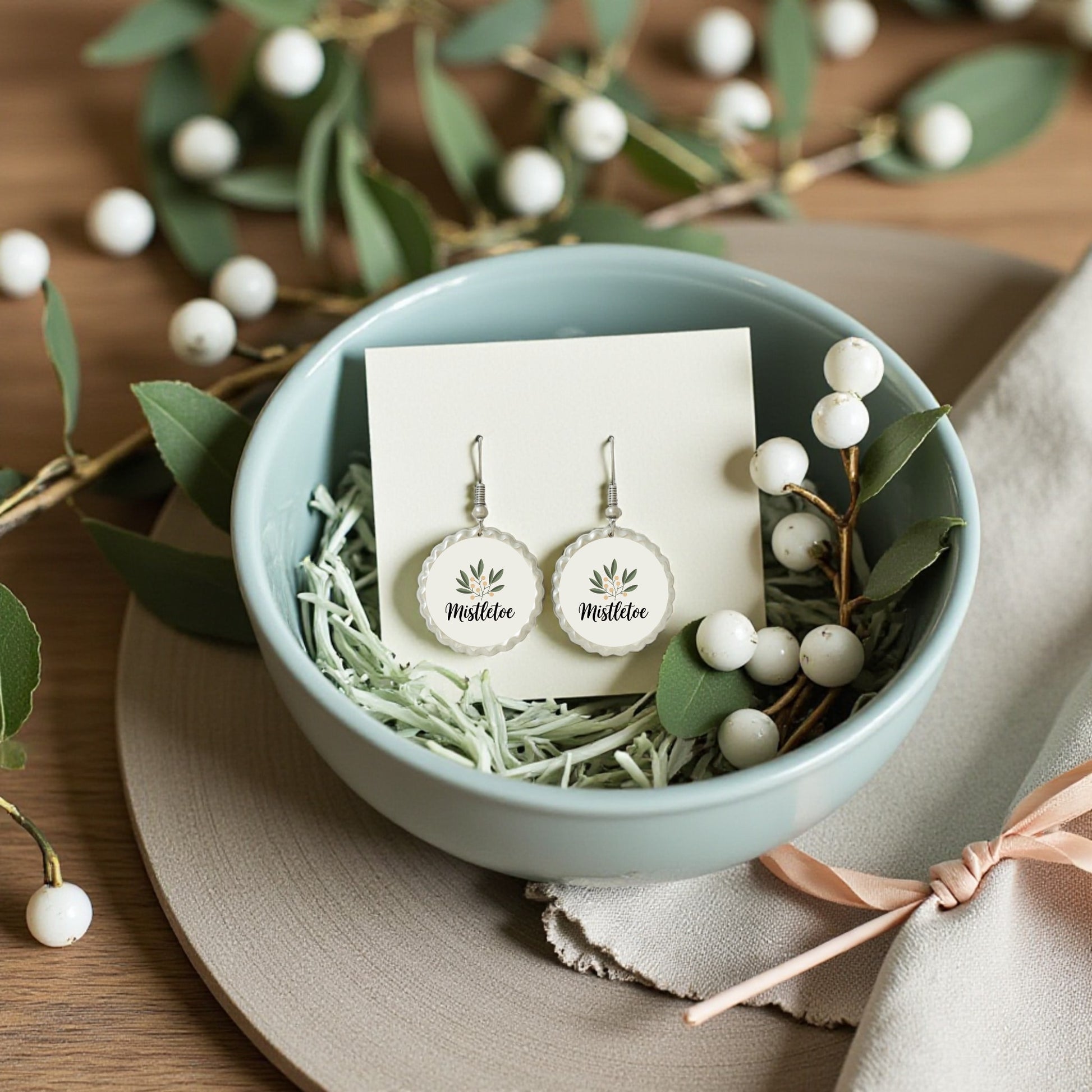 A pair of white round "Mistletoe Holiday Earrings | Botanical Christmas Jewelry" by Mirabilia Boutique rest in a light blue bowl with shredded paper, surrounded by artificial mistletoe and white berries on a wooden surface.