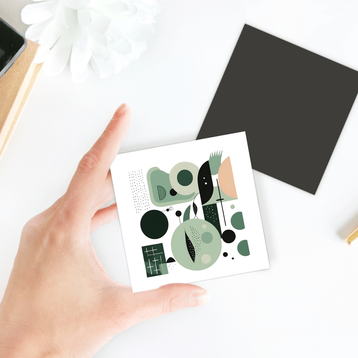 A hand holds the Spring Geometry Mid Century Modern Magnet by Mirabilia Boutique, featuring abstract geometric art in green, black, and peach tones. Nearby are a black card, white flower, and part of a notebook on a white surface.