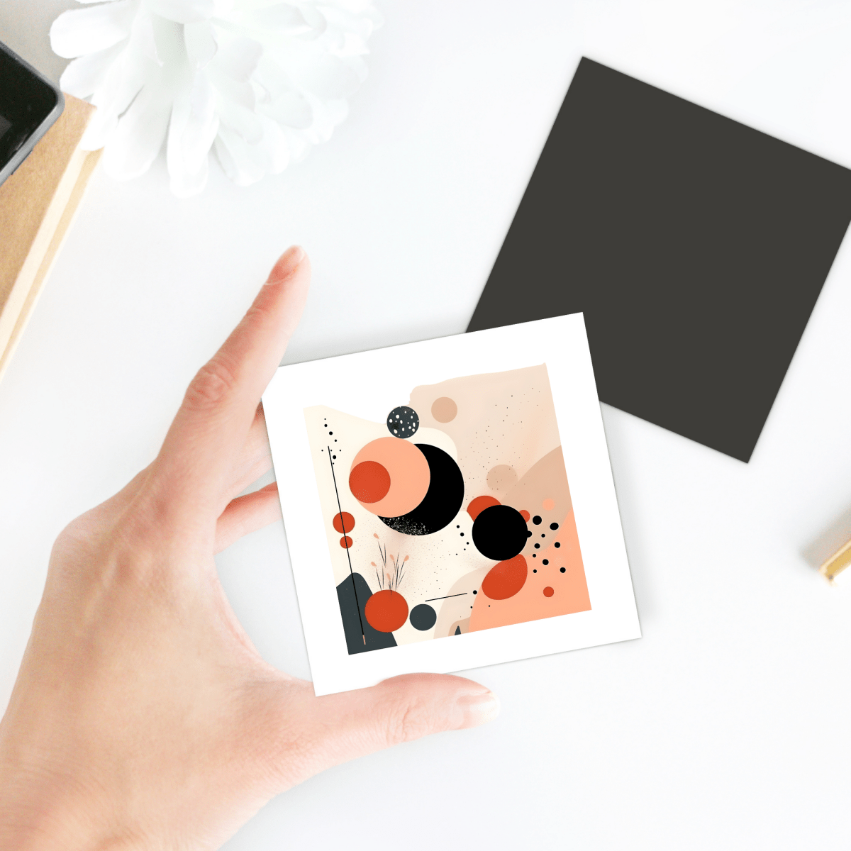 A hand holds the Solar Bloom Mid Century Modern Magnet by Mirabilia Boutique, featuring bold abstract shapes in black, orange, and beige. A black card, white flower, and desk items are visible in the background.