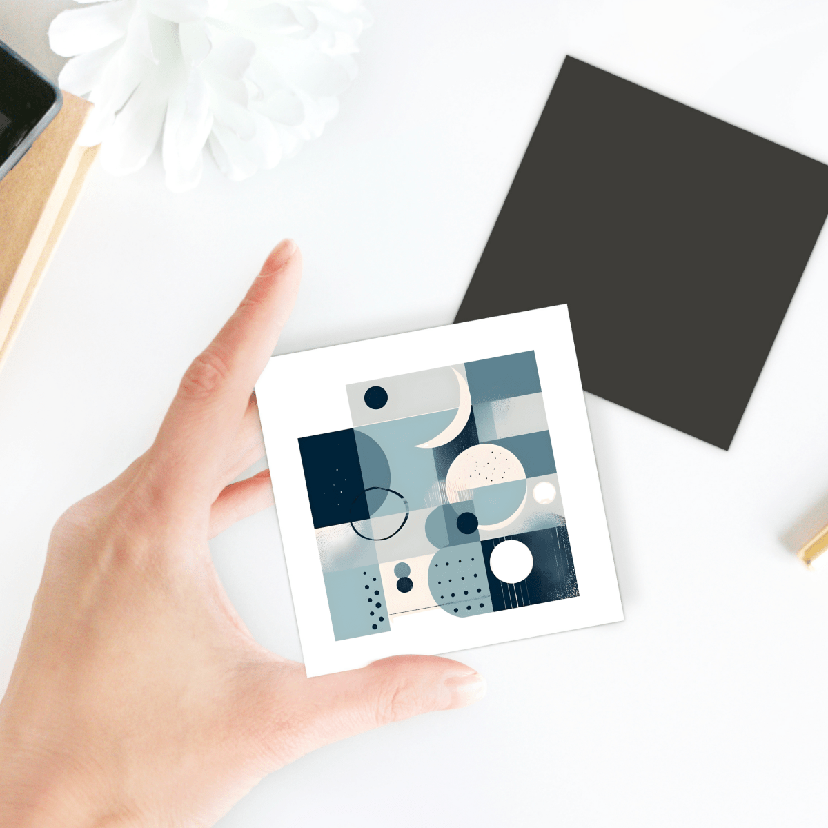 A hand holds the Moonlit Geometry Mid Century Modern Magnet by Mirabilia Boutique—a small square magnet with abstract blue, white, and beige shapes; nearby rest a black square accent and a white flower on a white surface.