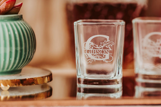 A Personalized Monogram Shot Glass from Mirabilia Boutique, custom engraved and clear, stands beside a green ribbed vase with a gold-rimmed base and a succulent on a reflective surface against a softly blurred background.