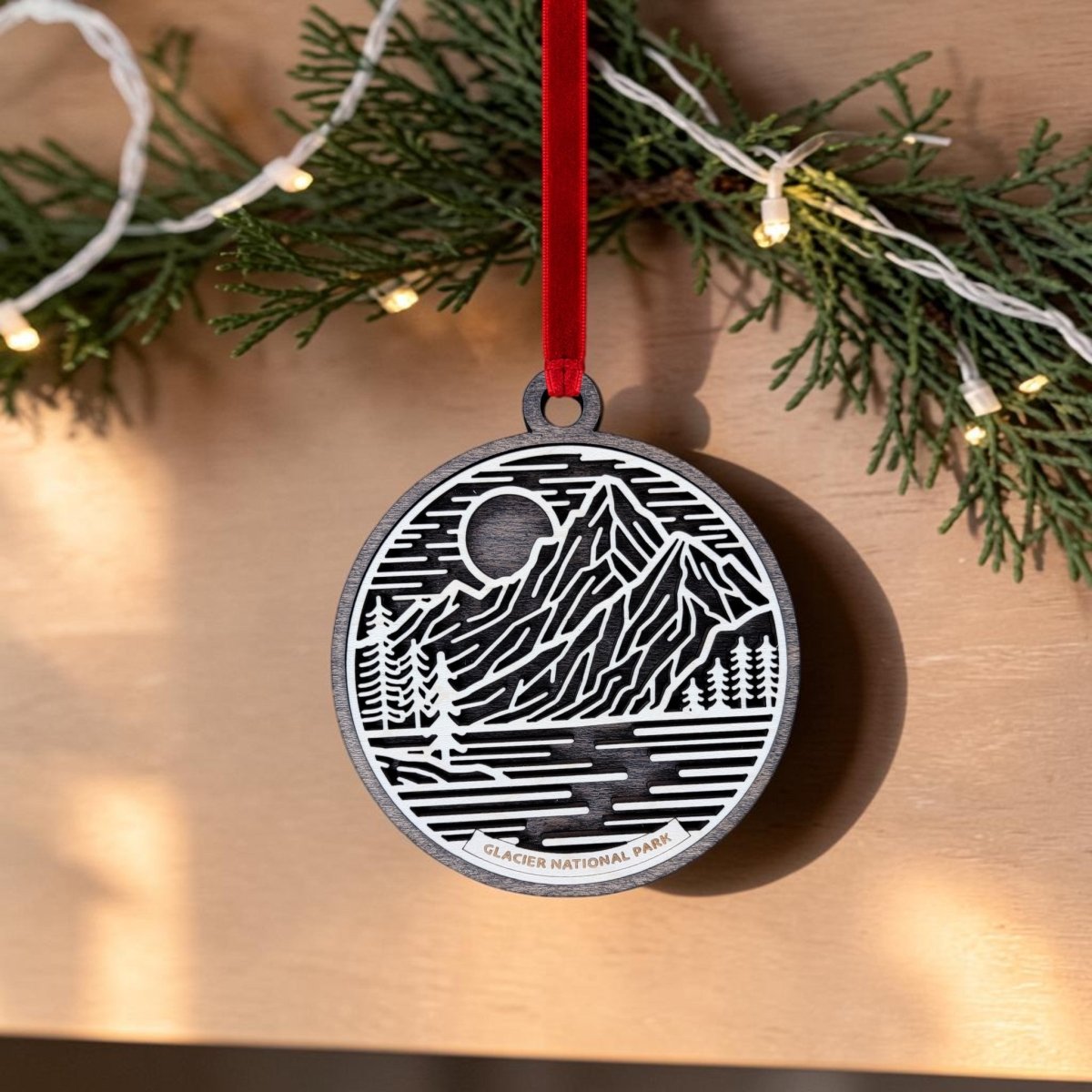 The Glacier National Park Line Art Christmas Ornament by Mirabilia Boutique displays an elegant line art design of a mountain, river, and trees with the words "Glacier National Park" and hangs from a red ribbon amid pine branches and string lights.