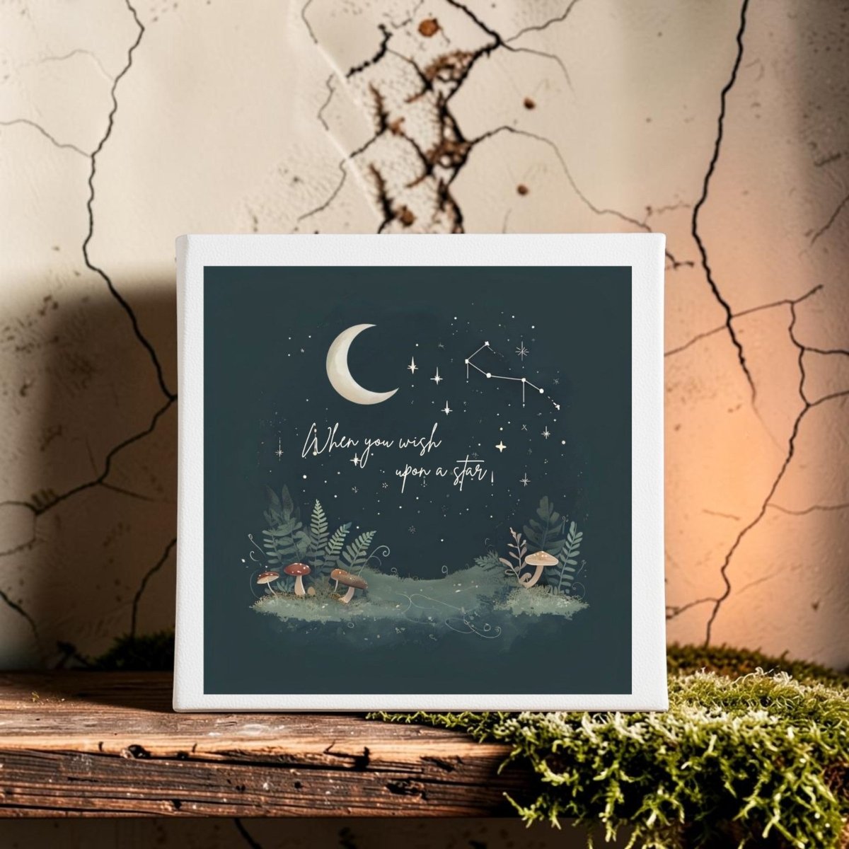 The When You Wish Upon a Star Woodland Canvas by Mirabilia Boutique displays a dreamy scene with a crescent moon, twinkling stars, and enchanting mushrooms on a dark backdrop. Featuring the saying “When you wish upon a star,” this piece is ideal as moon and stars decor. Shown resting on a wooden surface near moss and little plants against a cracked wall.