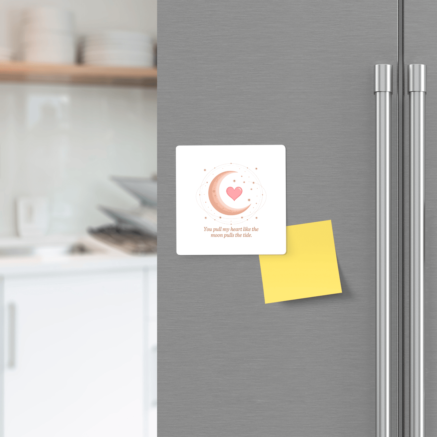 A stainless steel fridge displays two magnets: Mirabilia Boutique's Moon and Heart Love Quote Magnet holds a white card with a pink heart and moon, while another secures a blank yellow sticky note. Kitchen shelves and dishes appear in the background.