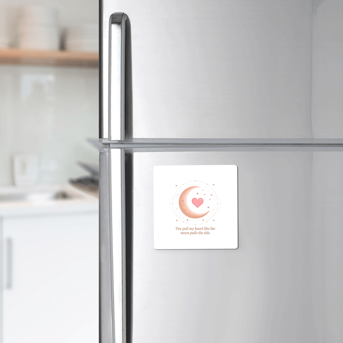 A silver fridge displays the Mirabilia Boutique Moon and Heart Love Quote Magnet—a square celestial magnet with a crescent moon, heart, and quote—in a modern kitchen with shelves and dishes in the background.