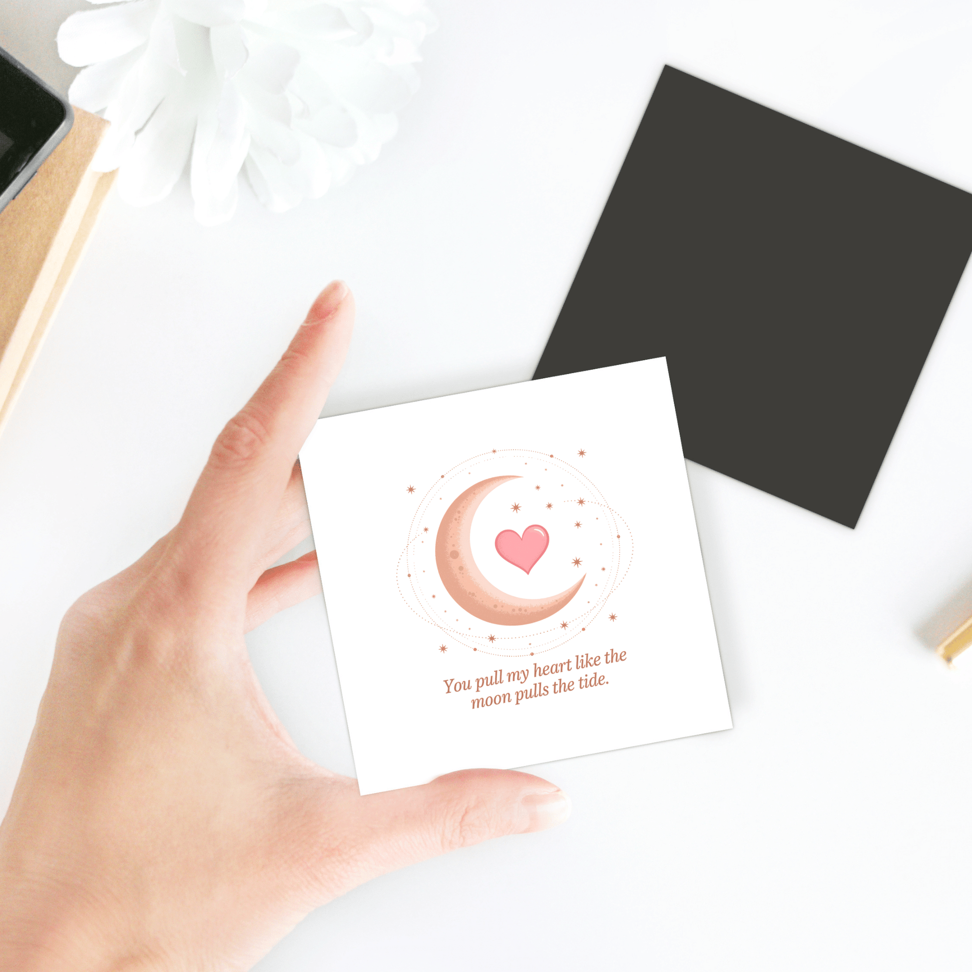 A hand holds the Moon and Heart Love Quote Magnet by Mirabilia Boutique, featuring a crescent moon, heart, and stars with the words, "You pull my heart like the moon pulls the tide." Other items are blurred in the background.