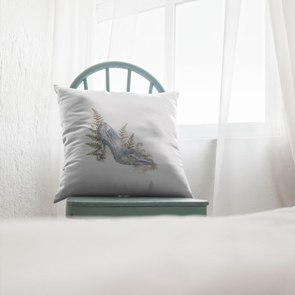 An Enchanted Glass Slipper Throw Pillow by Mirabilia Boutique, adorned with an illustration of a high-heeled shoe and ferns, rests on a light blue wooden chair in front of a window draped with sheer white curtains, bringing a touch of fairytale charm to the home décor.