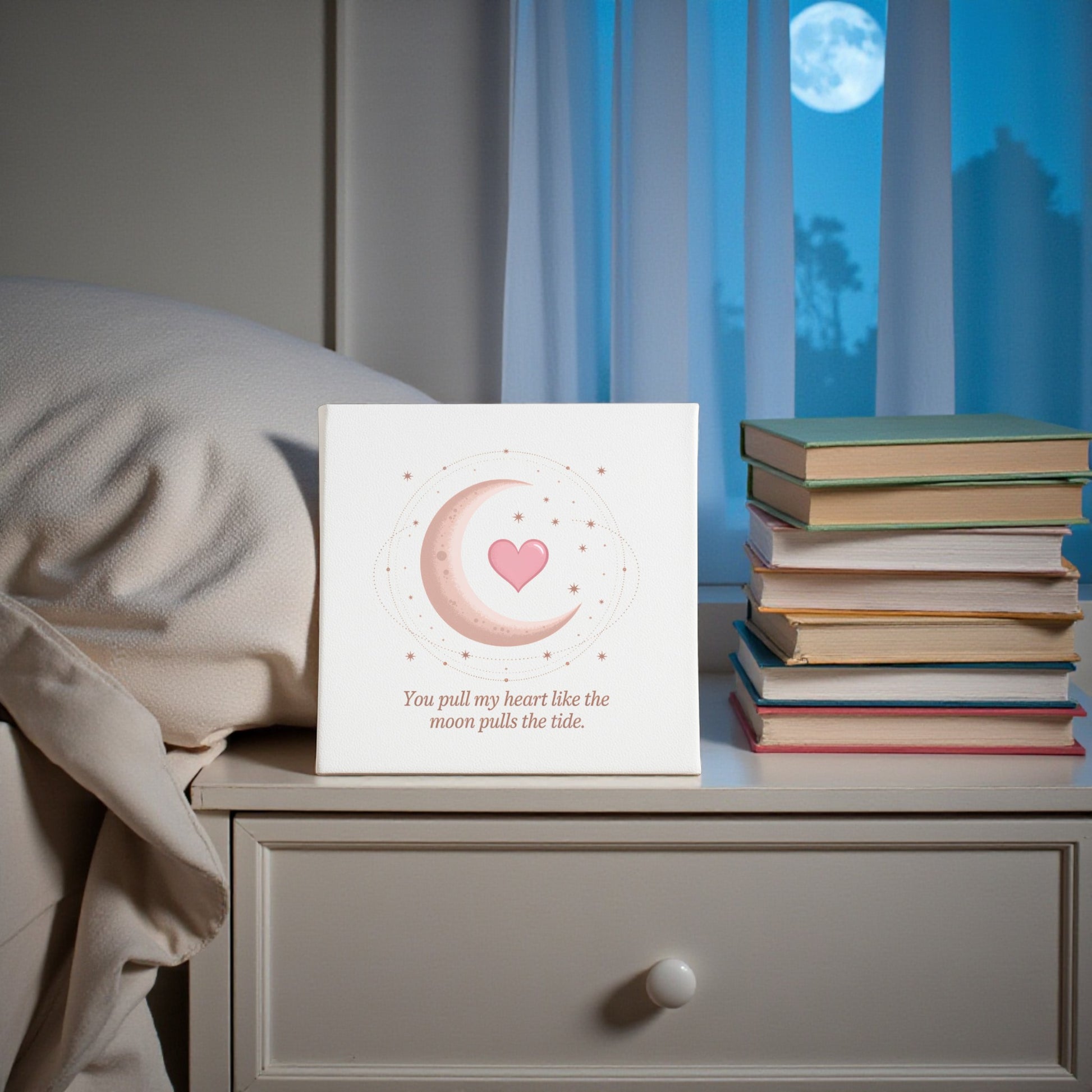The "Moon Pulls the Tide Love Quote Canvas" by Mirabilia Boutique rests on a bedside table with stacked books, while a full moon glows through the window—adding a romantic, celestial touch to your home decor.