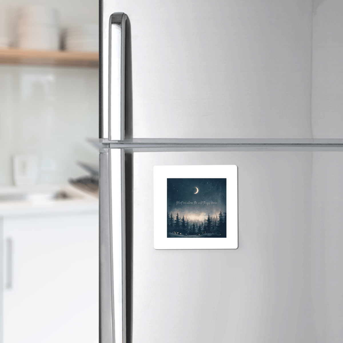 A silver refrigerator is accented with the Meet Me Where the Wild Things Dream Magnet from Mirabilia Boutique—a nature-inspired square magnet featuring a night forest scene, crescent moon, and stars. This celestial decor pops against white shelves and dishes, with a softly blurred kitchen counter in the background.