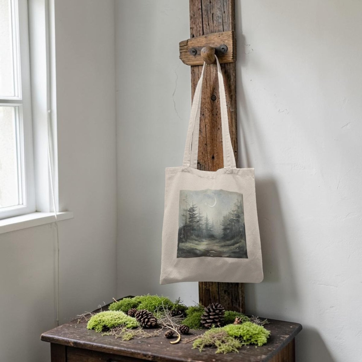 The "Some Paths Are Meant to Be Wandered" Tote Bag by Mirabilia Boutique, featuring a forest and moon design in beige, hangs from a wooden hook above a wooden table adorned with pinecones and moss in a softly lit room.