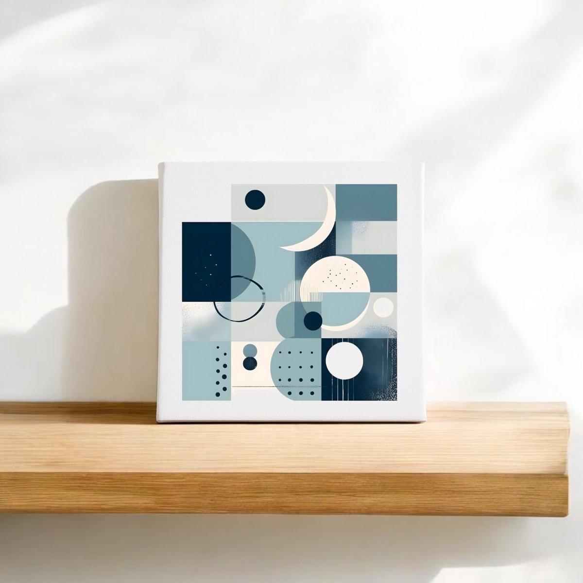 The Moonlit Geometry Mid Century Modern Canvas by Mirabilia Boutique features abstract blue, white, and beige shapes and rests on a light wooden shelf against a white wall, illuminated by soft sunlight and gentle shadows.