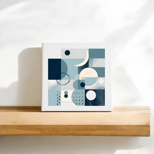 The Moonlit Geometry Mid Century Modern Canvas by Mirabilia Boutique features abstract blue, white, and beige shapes and rests on a light wooden shelf against a white wall, illuminated by soft sunlight and gentle shadows.