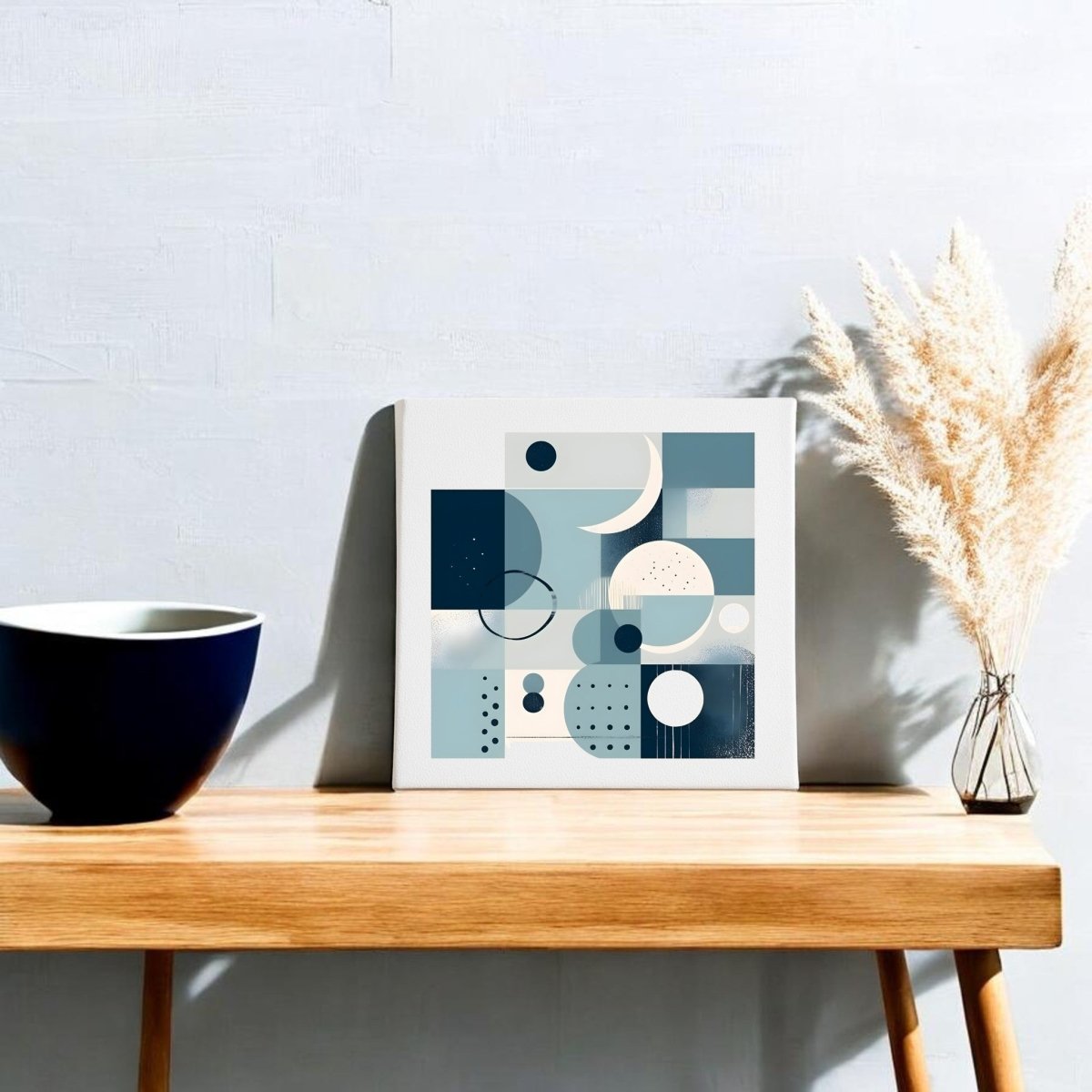 A wooden table holds a blue and white bowl, a glass vase with dried beige pampas grass, and the Moonlit Geometry Mid Century Modern Canvas by Mirabilia Boutique, featuring abstract blue and cream art, set against a light gray wall.