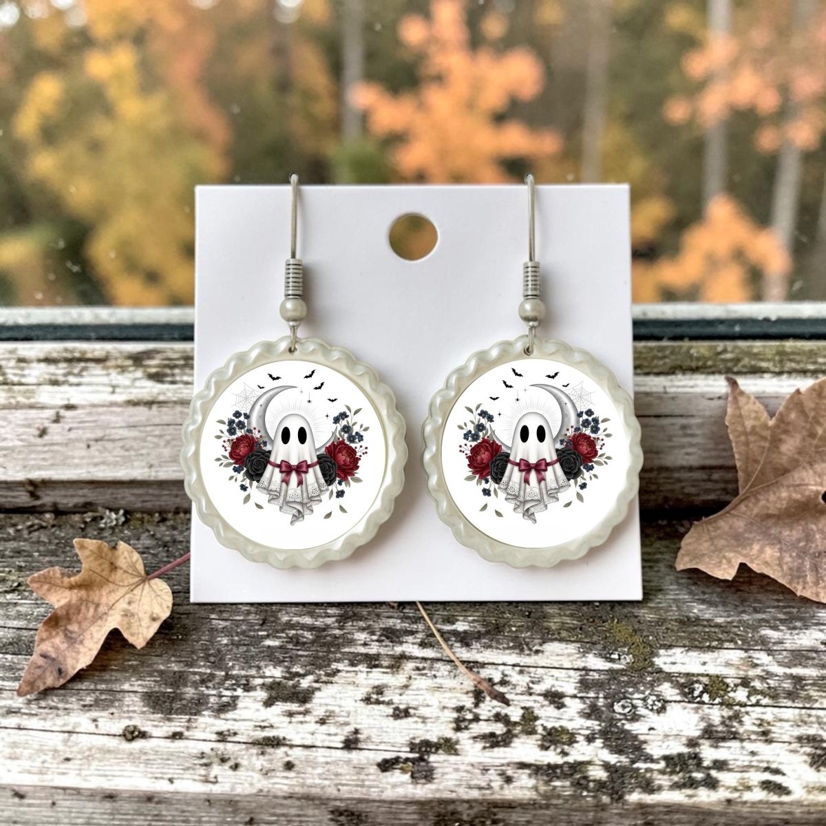 The Crescent Moon Lace Ghost Scalloped Earrings by Mirabilia Boutique feature a round, lace design with whimsical ghosts, red roses, and black leaves. Displayed elegantly on a card with autumn leaves and a softly blurred fall backdrop, these charming vintage-inspired earrings are perfect for the fall season.