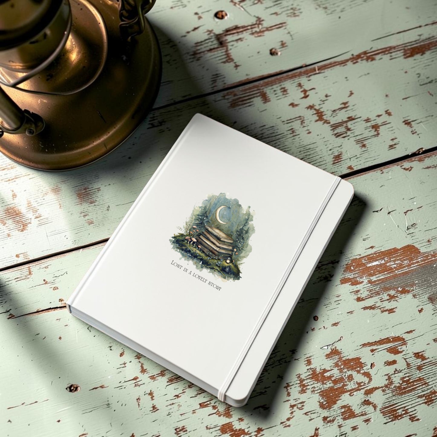 A white hardcover “Lost in a Lovely Story Journal” from Mirabilia Boutique, featuring a moon and book illustration with the phrase “Lost in a lovely story,” rests on a distressed green wooden table beside a brass lamp, evoking the charm of a storybook journal.