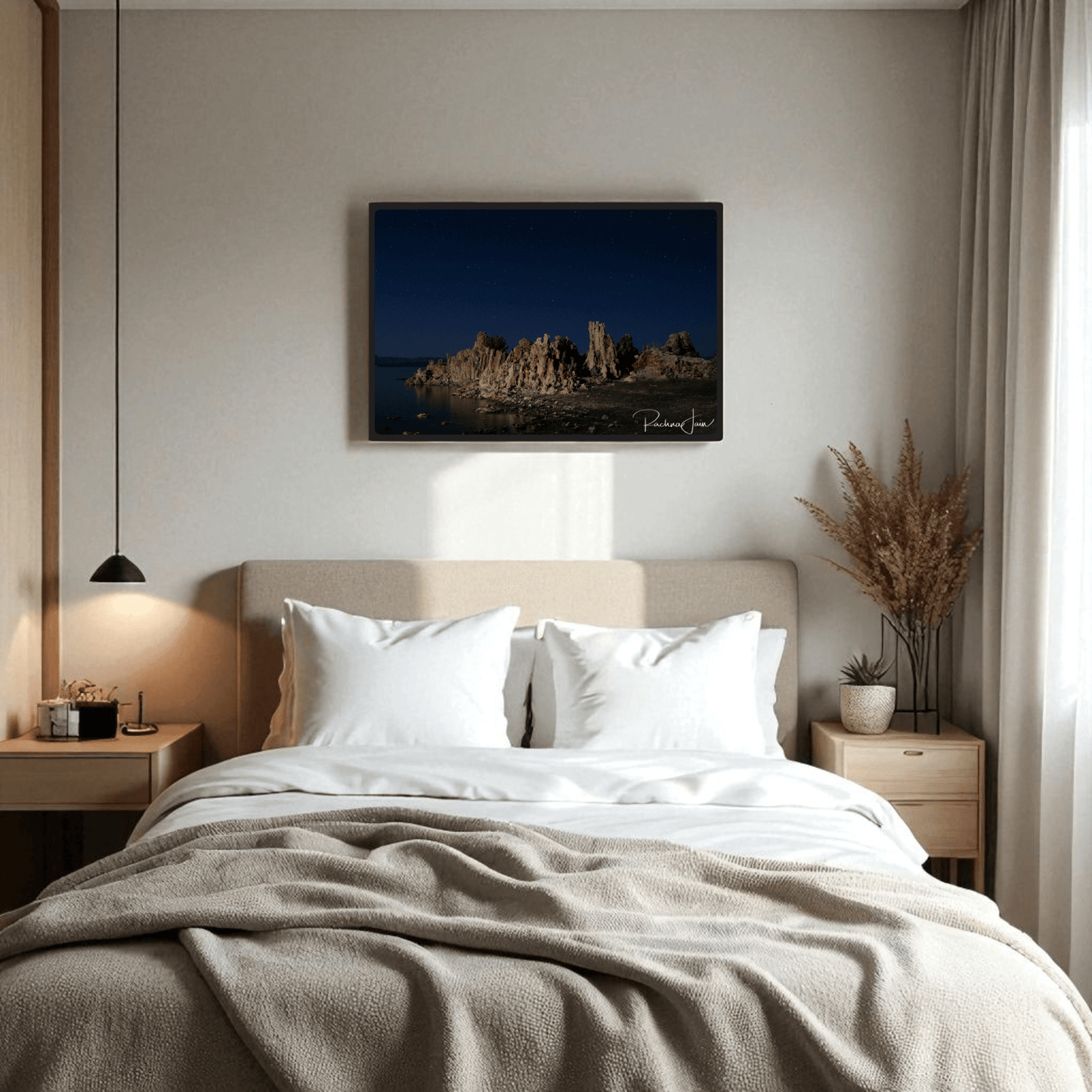 A cozy bedroom features a beige bed with white pillows and a blanket. Two nightstands with lamps add warmth, while a vase of dried plants sits on one side. Above the bed hangs "Moonlit Tufas at Mono Lake," a Mirabilia Boutique framed art print by Rachna Jain.