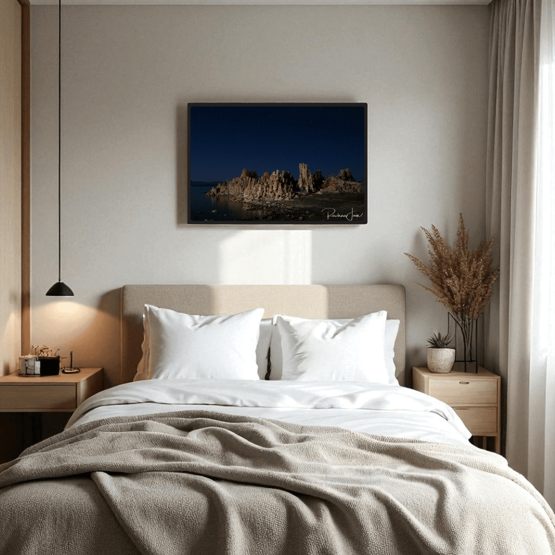 A cozy bedroom features a beige bed with white pillows and a blanket. Two nightstands with lamps add warmth, while a vase of dried plants sits on one side. Above the bed hangs "Moonlit Tufas at Mono Lake," a Mirabilia Boutique framed art print by Rachna Jain.