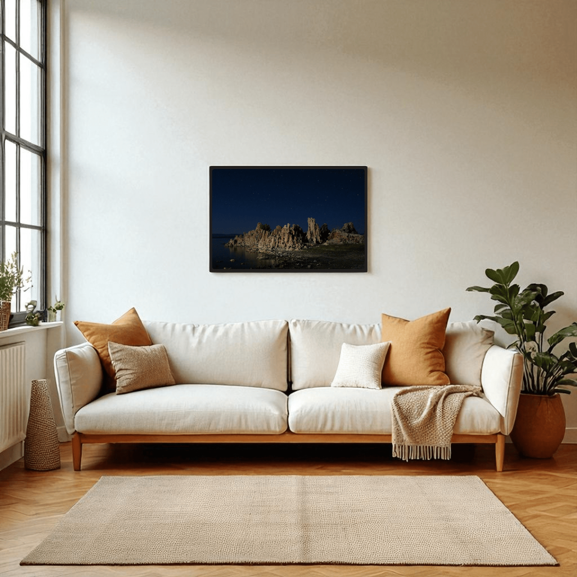 A modern living room with a white sofa, beige and orange cushions, a throw, green plants, large window, and nature photography—a Mirabilia Boutique "Moonlit Tufas at Mono Lake" framed art print by Rachna Jain above the sofa.