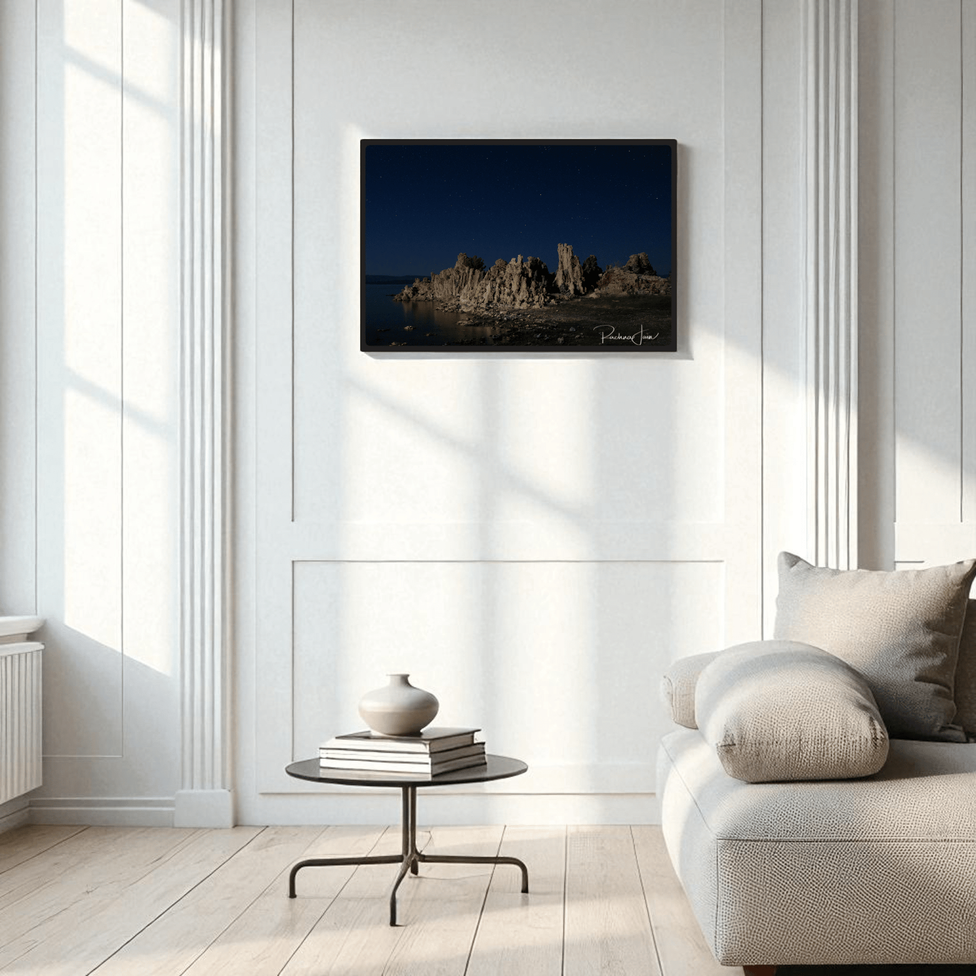 A modern, minimalist living room with white walls, a cozy beige sofa, a round black coffee table, and the "Moonlit Tufas at Mono Lake" framed art print by Rachna Jain from Mirabilia Boutique as fine art wall decor.