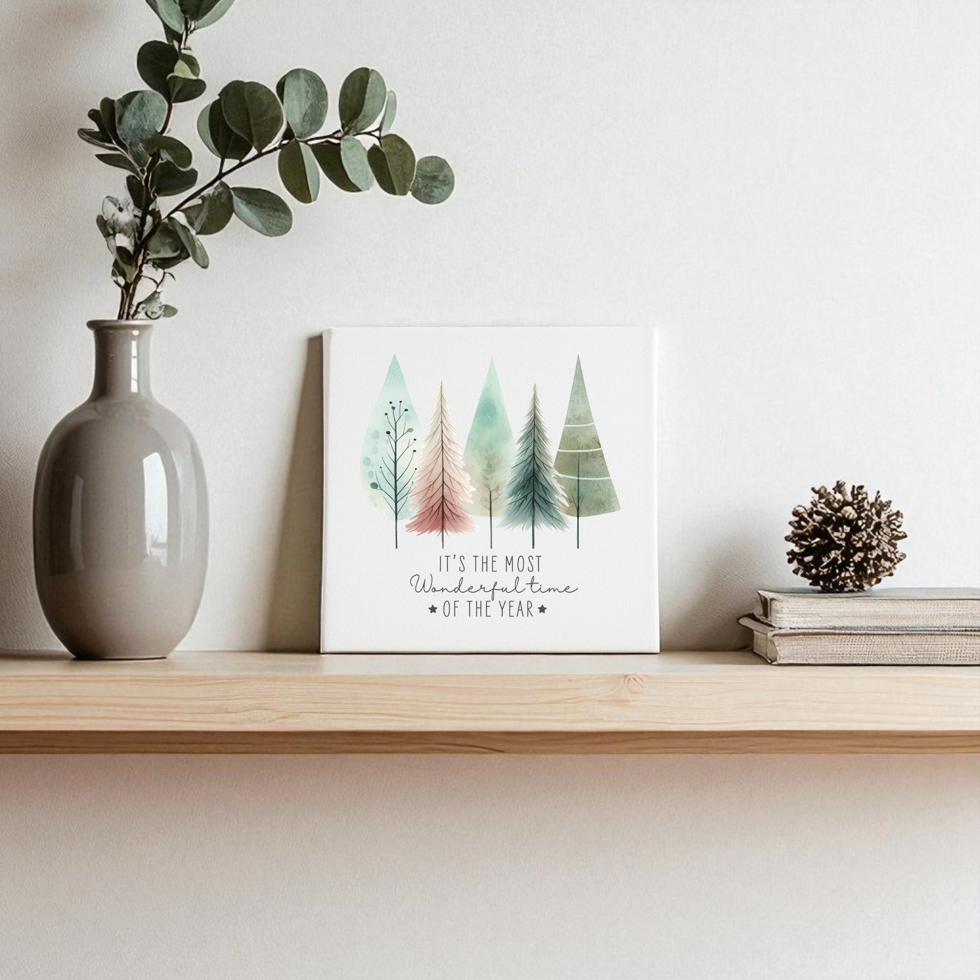 A wooden shelf with winter decor: a gray vase with green branches, a pine cone, two stacked books, and the Mirabilia Boutique "Most Wonderful Time of Year Canvas | Pastel Watercolor Trees" against a light wall.