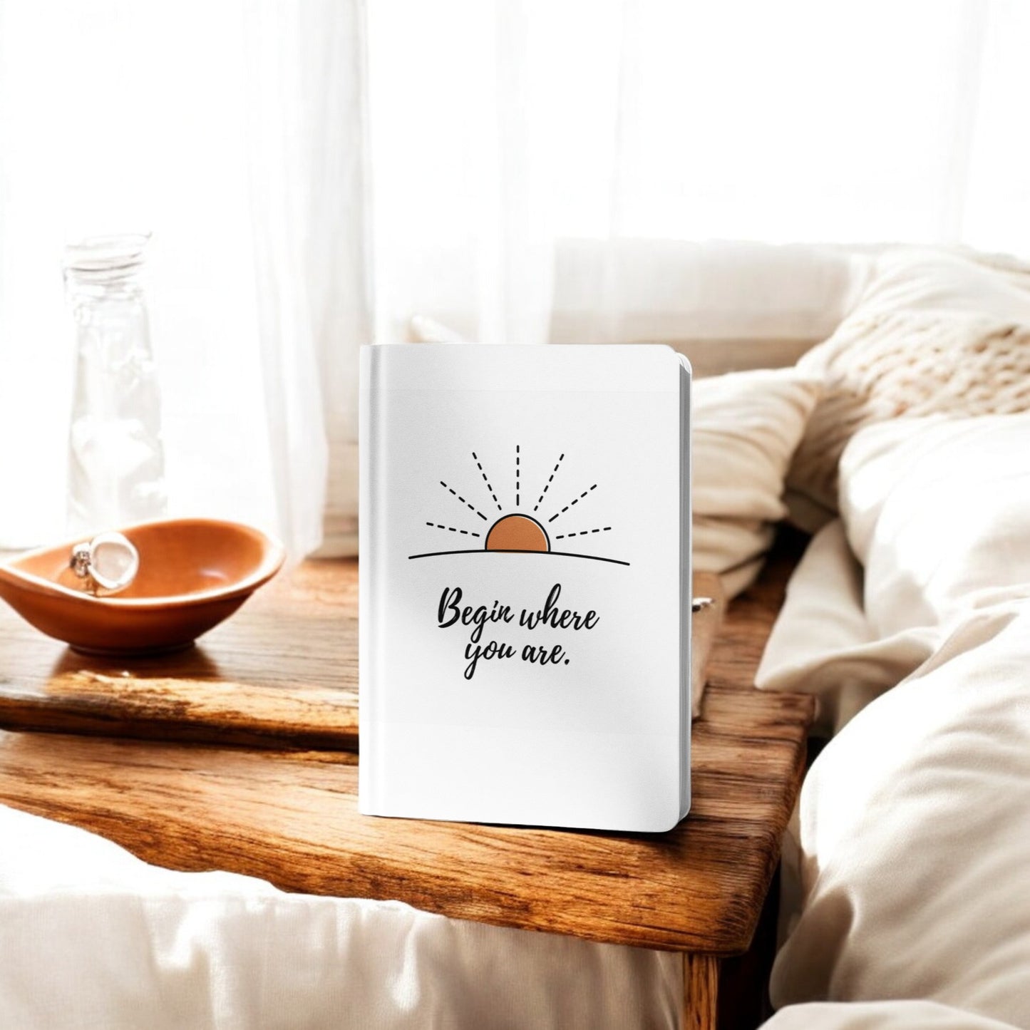 The Begin Where You Are Journal – Sunrise Quote Notebook by Mirabilia Boutique rests on a wooden tray by the bed, paired with a glass water bottle and brown dish. Bright natural light creates an inspiring space for morning journaling.
