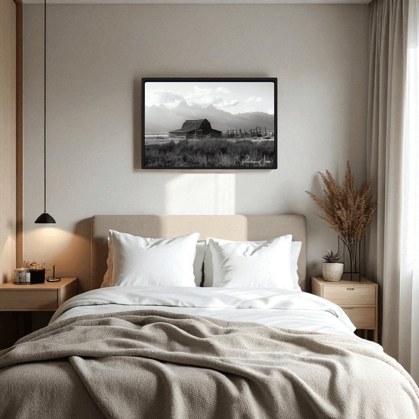 A cozy bedroom features a beige bed with white pillows and the Moulton Barn Grand Teton Black and White Framed Art Print by Rachna Jain from Mirabilia Boutique, elegantly displayed above the headboard. A nightstand with a lamp and decorative grass sits nearby.