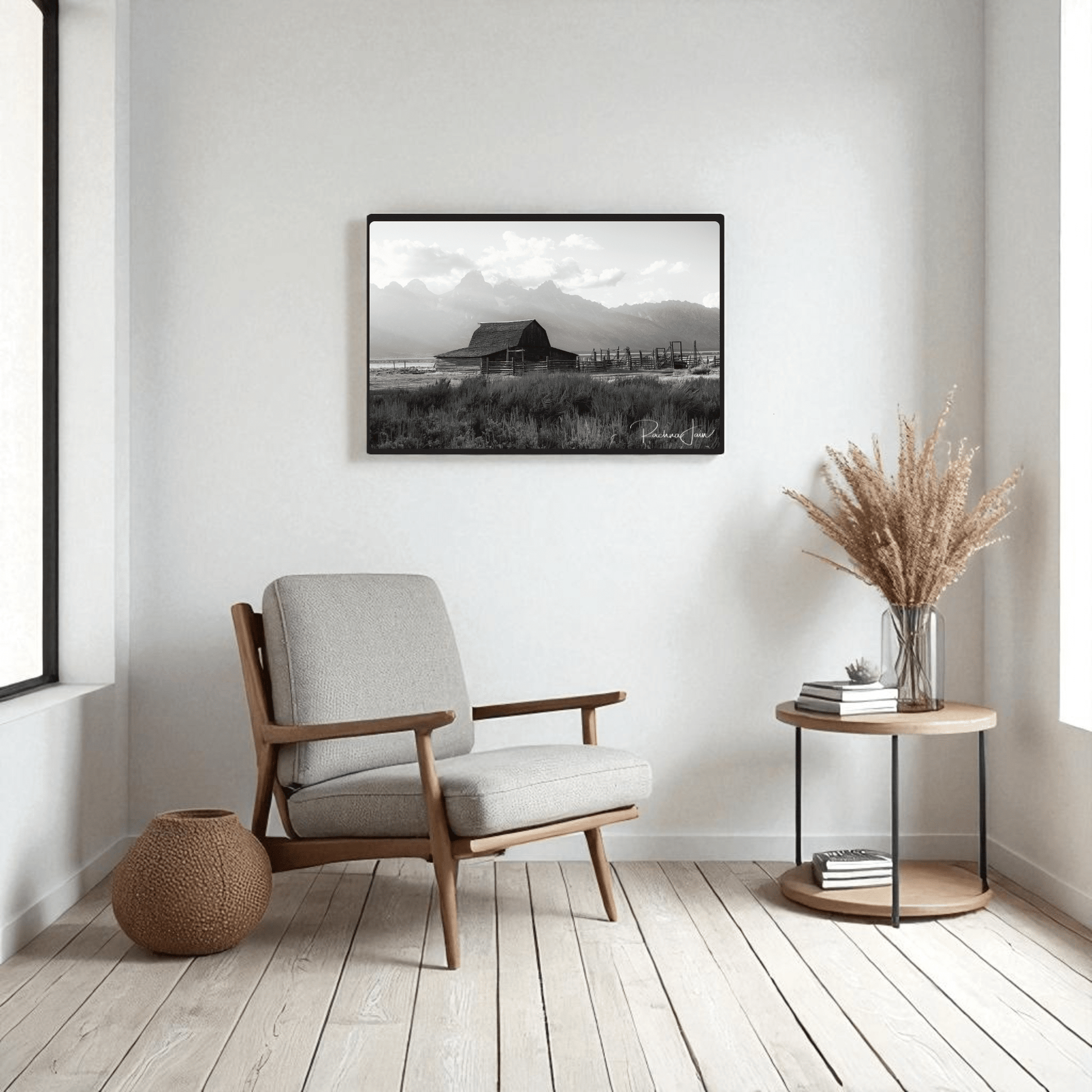 Minimalist living room with a light armchair, side table, vase of dried grasses, woven basket, and the Moulton Barn Grand Teton Black and White Framed Art Print by Rachna Jain from Mirabilia Boutique on the wall. Light wood floors and white walls complete the serene look.