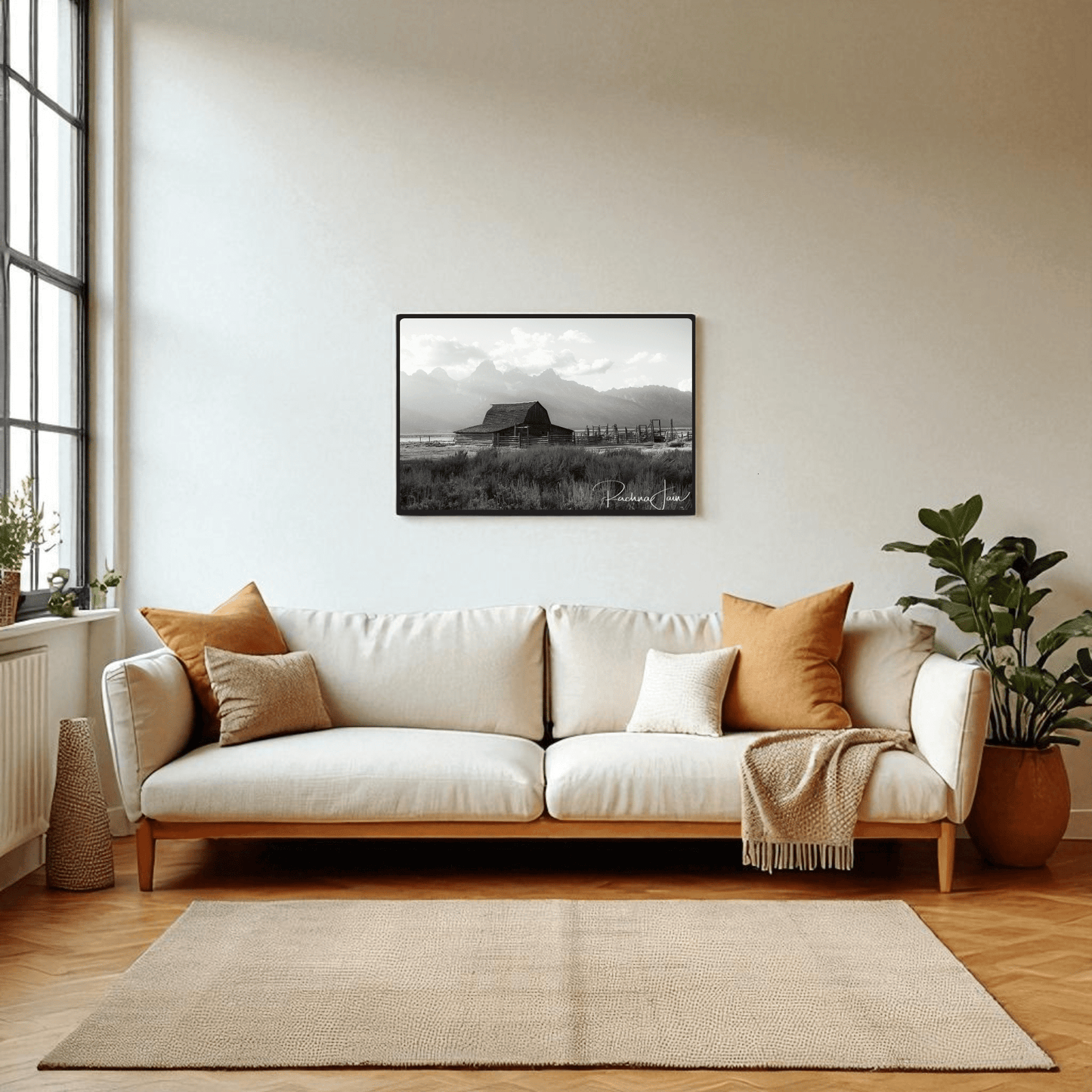 A modern living room with a white sofa, earth-toned pillows, potted plants, and a large window features the "Moulton Barn Grand Teton" black and white framed art print by Rachna Jain from Mirabilia Boutique above the sofa.