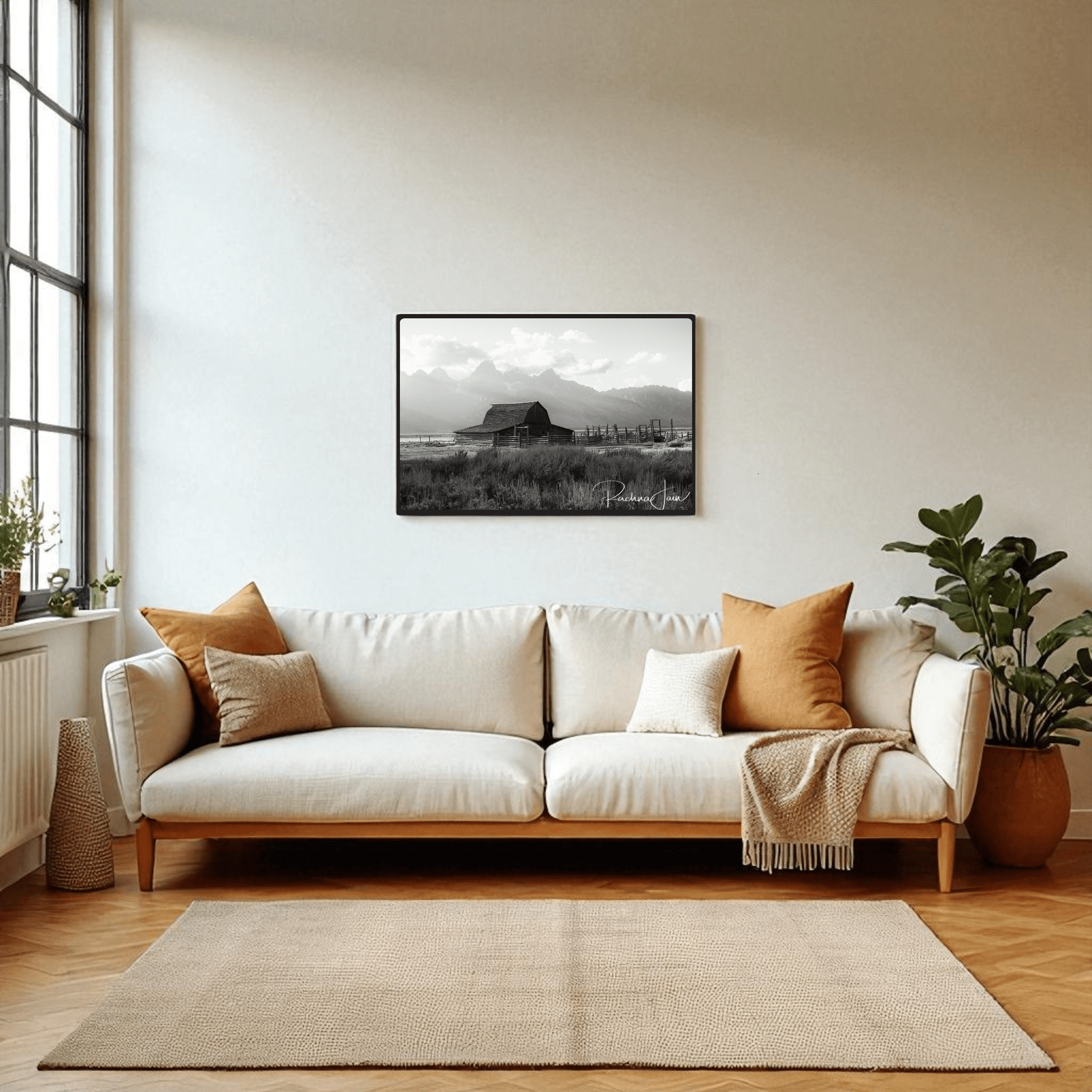 A modern living room with a white sofa, earth-toned pillows, potted plants, and a large window features the "Moulton Barn Grand Teton" black and white framed art print by Rachna Jain from Mirabilia Boutique above the sofa.