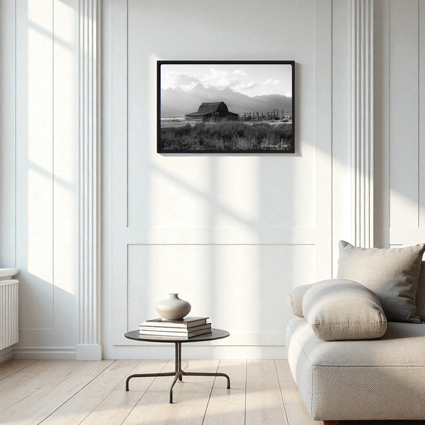 A modern living room with a light sofa, round glass coffee table with a vase, and the "Moulton Barn Grand Teton" black and white framed art print by Rachna Jain from Mirabilia Boutique. Natural light enhances the minimalist space.