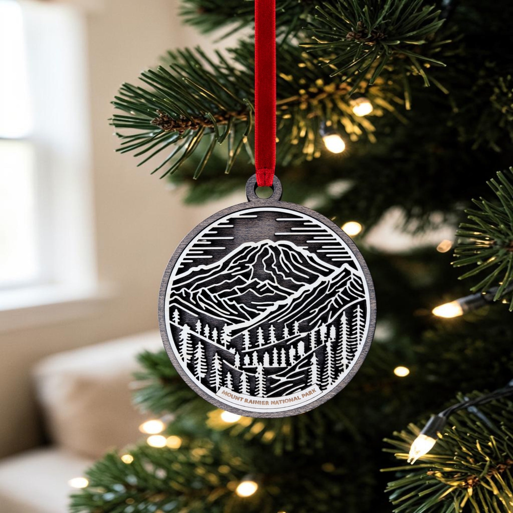 The Mount Rainier National Park Line Art Christmas Ornament by Mirabilia Boutique, crafted in an oval wooden design with a mountain and trees motif, hangs from a red ribbon amidst twinkling lights on a Christmas tree.