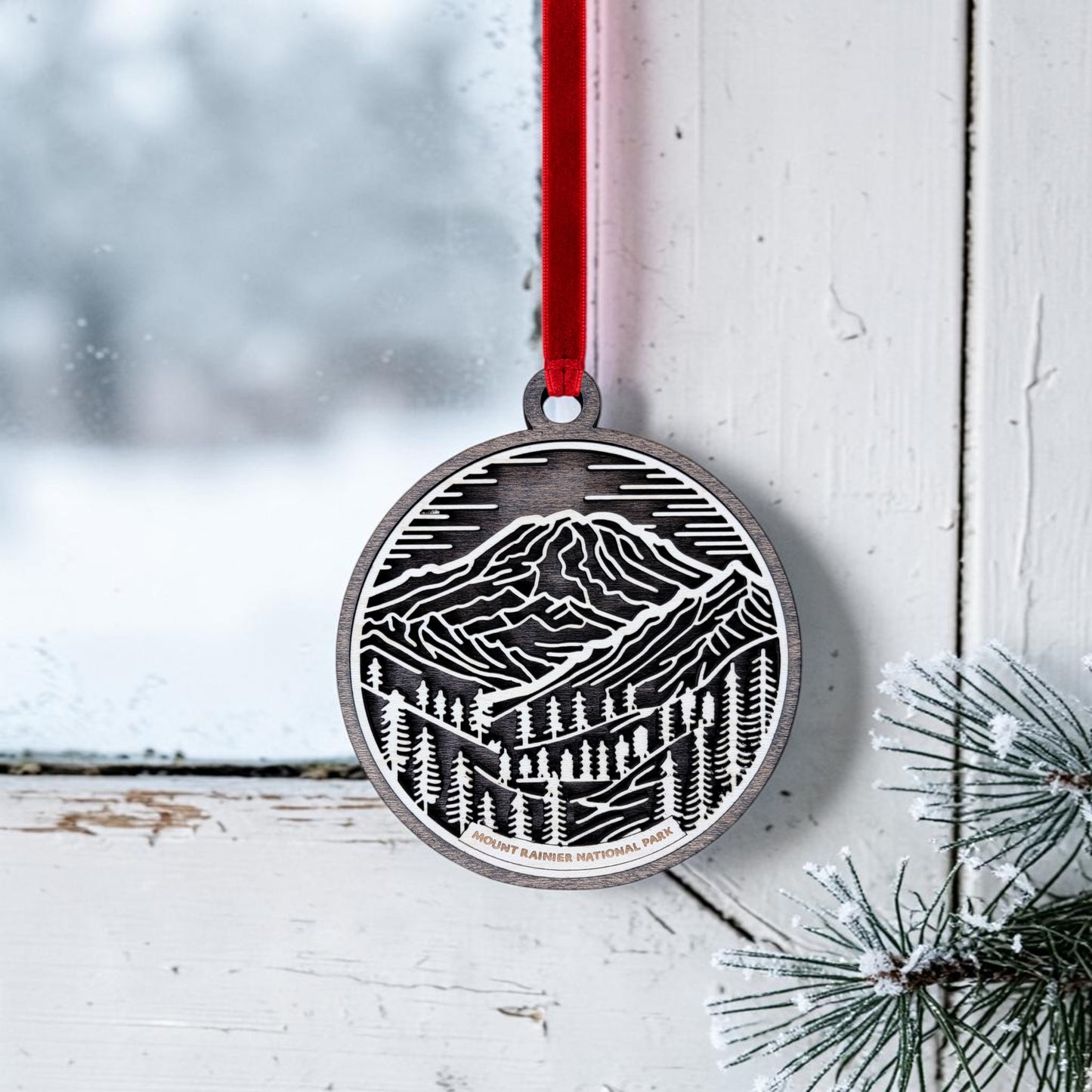 The Mount Rainier National Park Line Art Christmas Ornament by Mirabilia Boutique, featuring a black and white mountain and pine tree design on an oval wooden base, hangs from a red ribbon on a snowy window with pine branches visible in the corner.