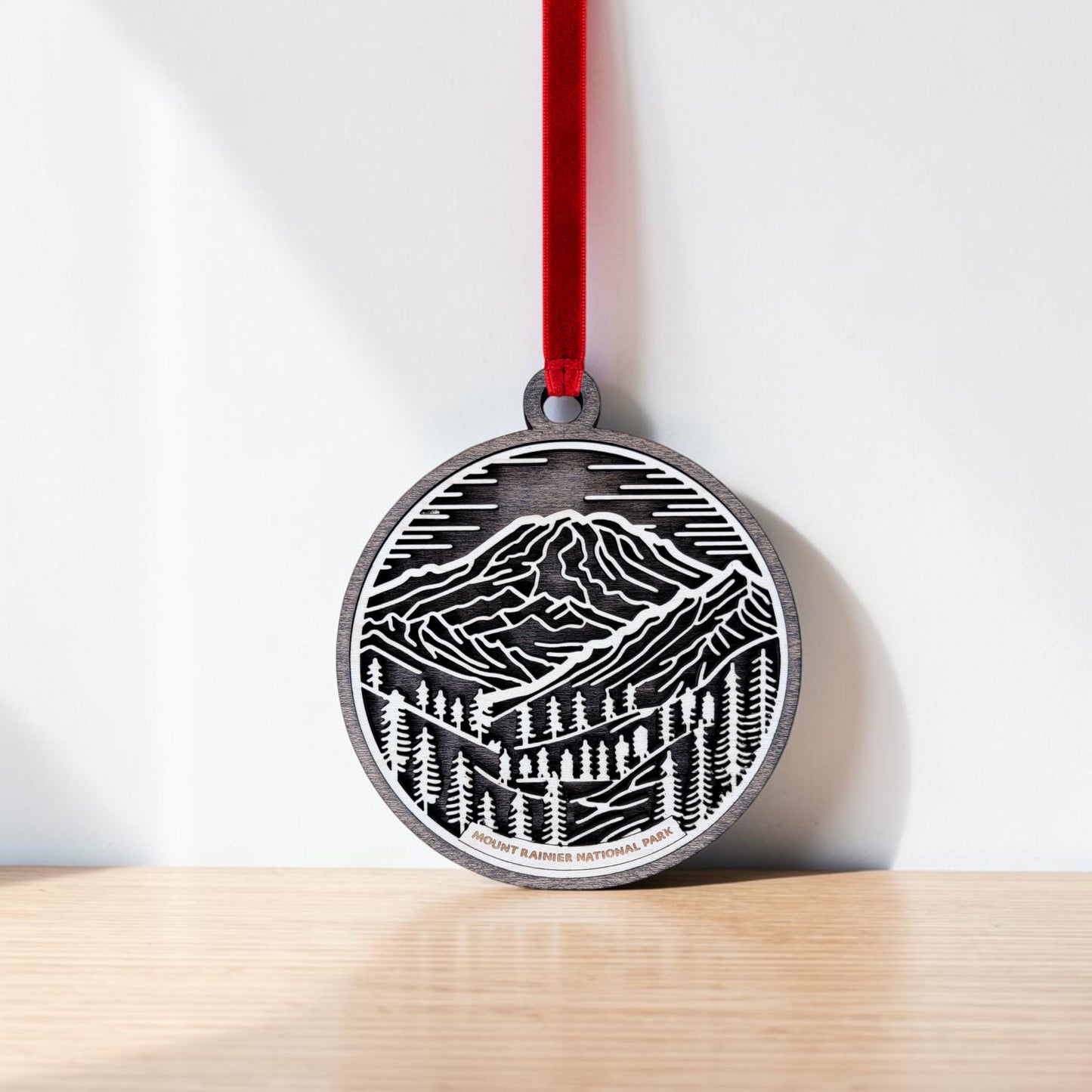 The Mount Rainier National Park Line Art Christmas Ornament by Mirabilia Boutique, featuring a round mountain and pine tree design, hangs from a red ribbon against a white wall and rests on a light wooden surface.