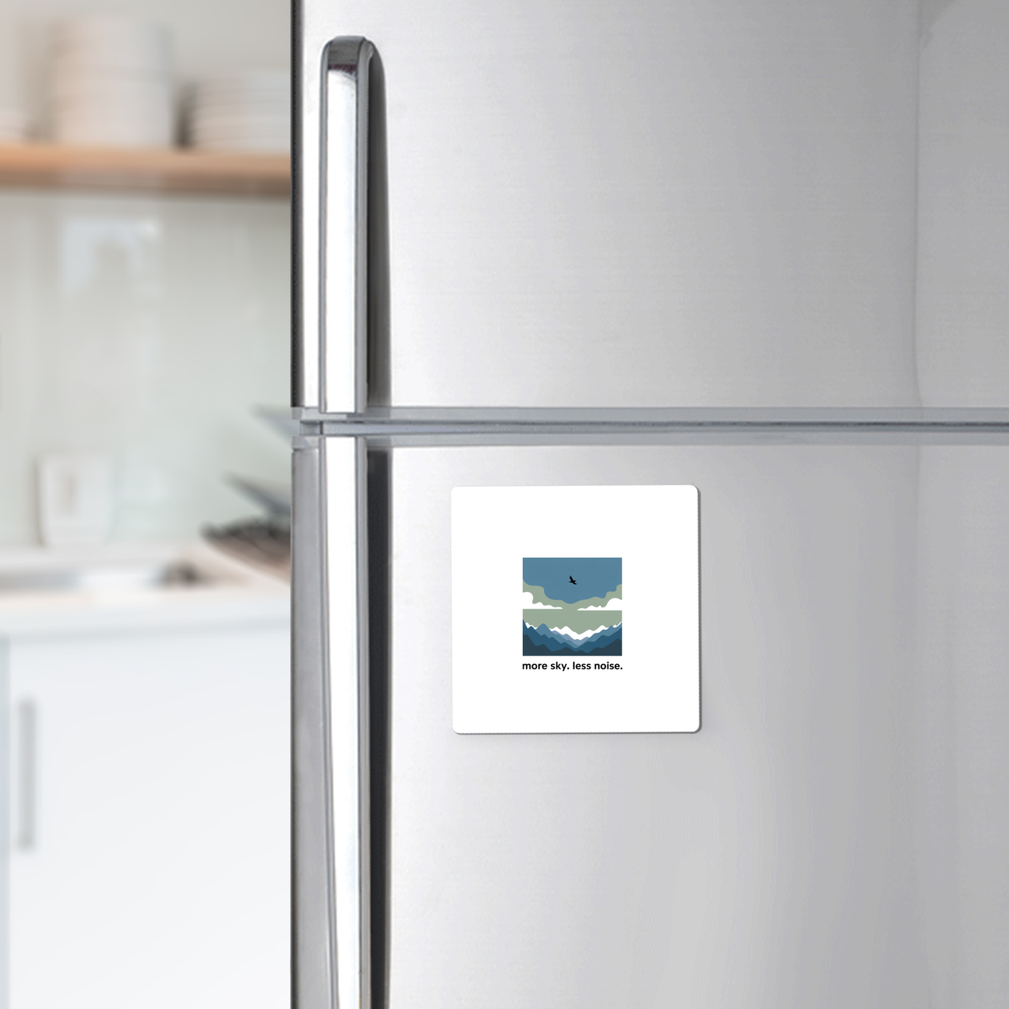 A silver fridge displays the “More Sky Less Noise Nature Magnet” by Mirabilia Boutique, featuring a minimalist landscape and bird. Ideal outdoor gift for minimalist decor lovers. Modern kitchen with white shelves in the background.