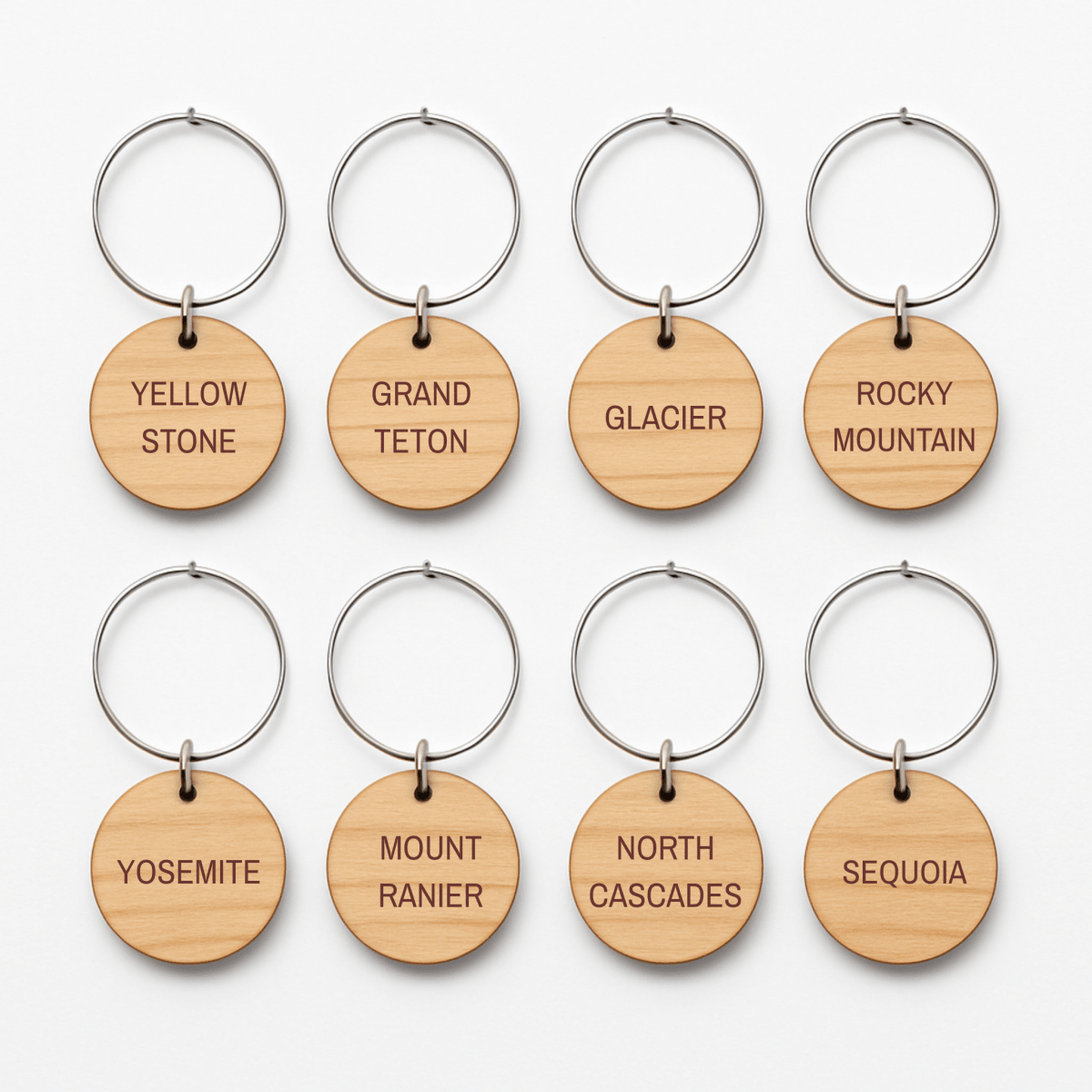 The Mountain Majesty National Park Wine Charms by Mirabilia Boutique feature eight round wooden keychains with metal rings, each engraved with a national park name—perfect for outdoor-inspired gatherings.