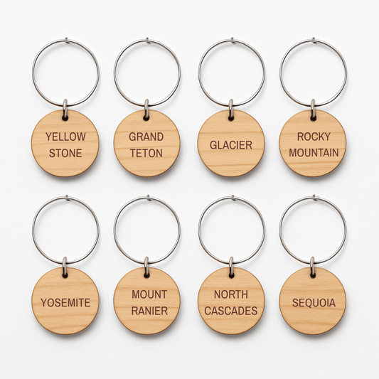 The Mountain Majesty National Park Wine Charms by Mirabilia Boutique feature eight round wooden keychains with metal rings, each engraved with a national park name—perfect for outdoor-inspired gatherings.