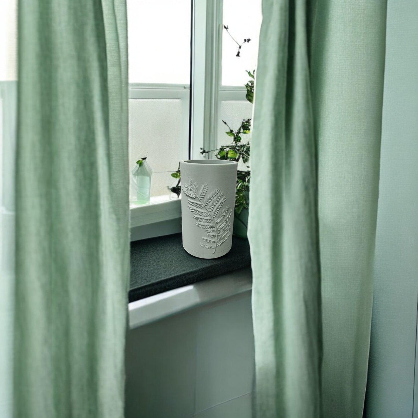 A Mirabilia Boutique Fern Emboss Vase in soft green stands on a windowsill between pale green curtains. Nearby, a small bottle and leafy plant enhance the minimalist decor, while soft light shines through the frosted glass.