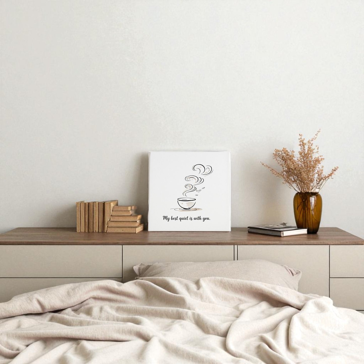 A minimal bedroom features an unmade bed facing a wooden dresser topped with books, a vase of dried flowers, and Mirabilia Boutique’s Best Quiet Canvas—a minimalist coffee quote wall art print.