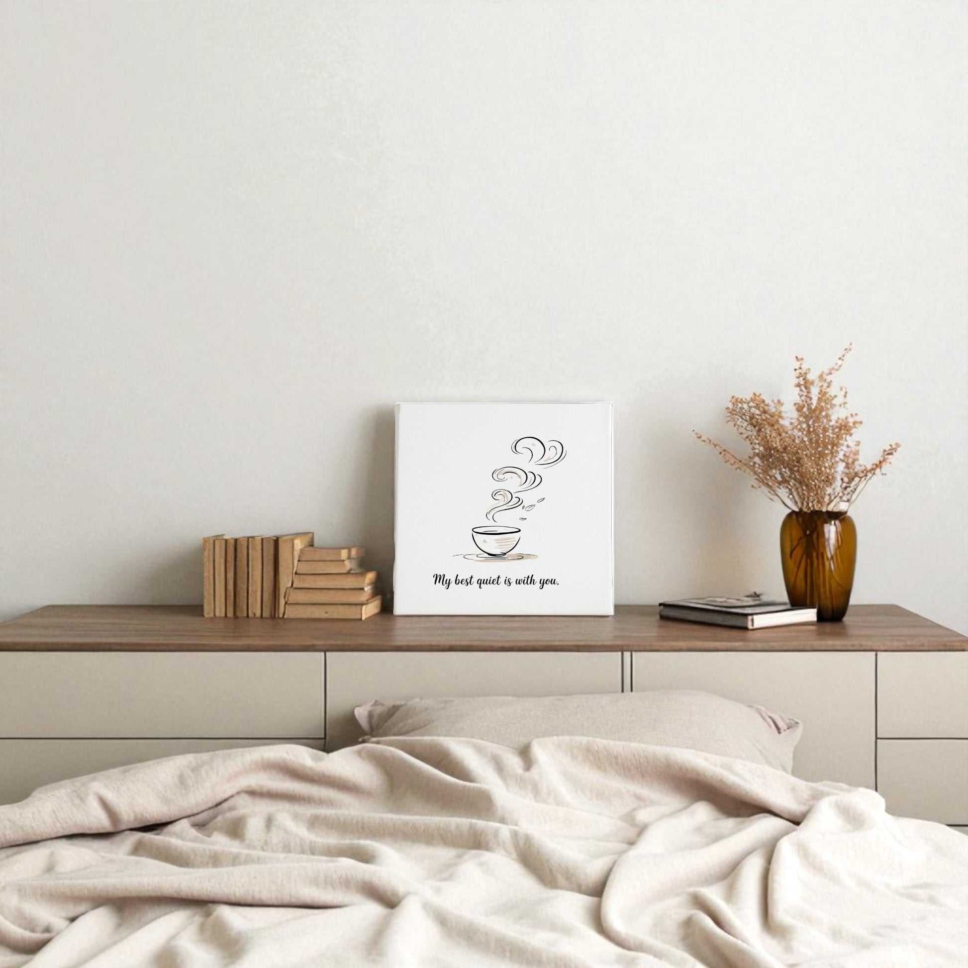 A minimal bedroom features an unmade bed facing a wooden dresser topped with books, a vase of dried flowers, and Mirabilia Boutique’s Best Quiet Canvas—a minimalist coffee quote wall art print.