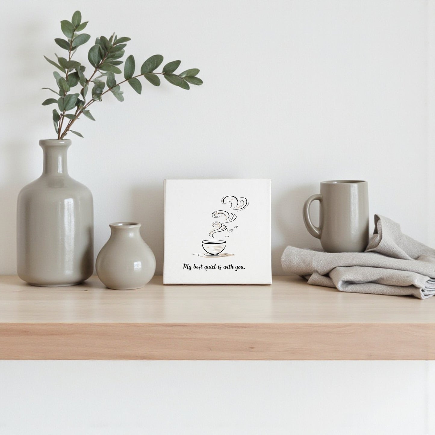 A wooden shelf holds neutral ceramic vases, a mug, folded cloth, and Mirabilia Boutique’s “Best Quiet Canvas”—a minimalist coffee quote wall art, ideal as a heartfelt gift or tranquil decor for your space.