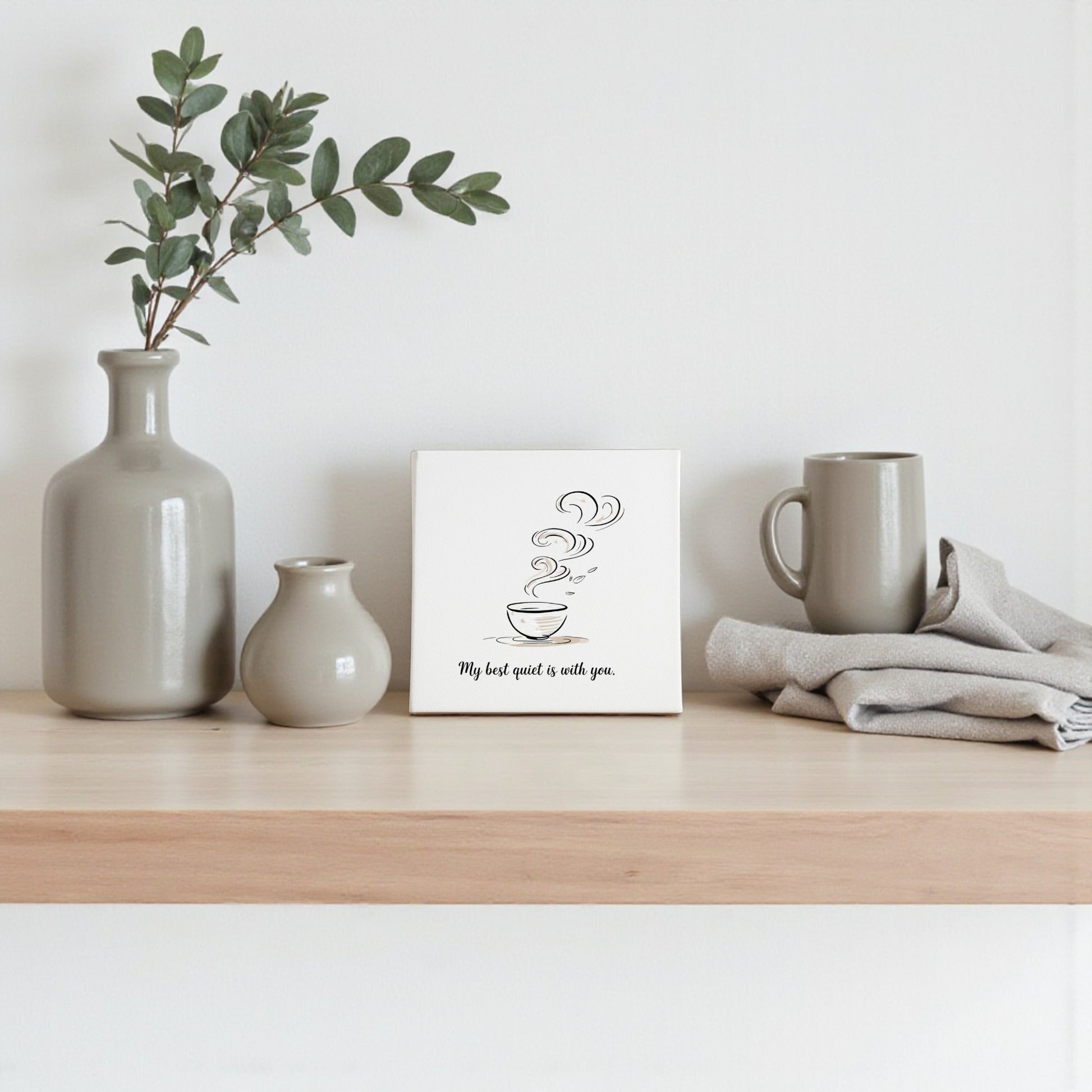A wooden shelf holds neutral ceramic vases, a mug, folded cloth, and Mirabilia Boutique’s “Best Quiet Canvas”—a minimalist coffee quote wall art, ideal as a heartfelt gift or tranquil decor for your space.