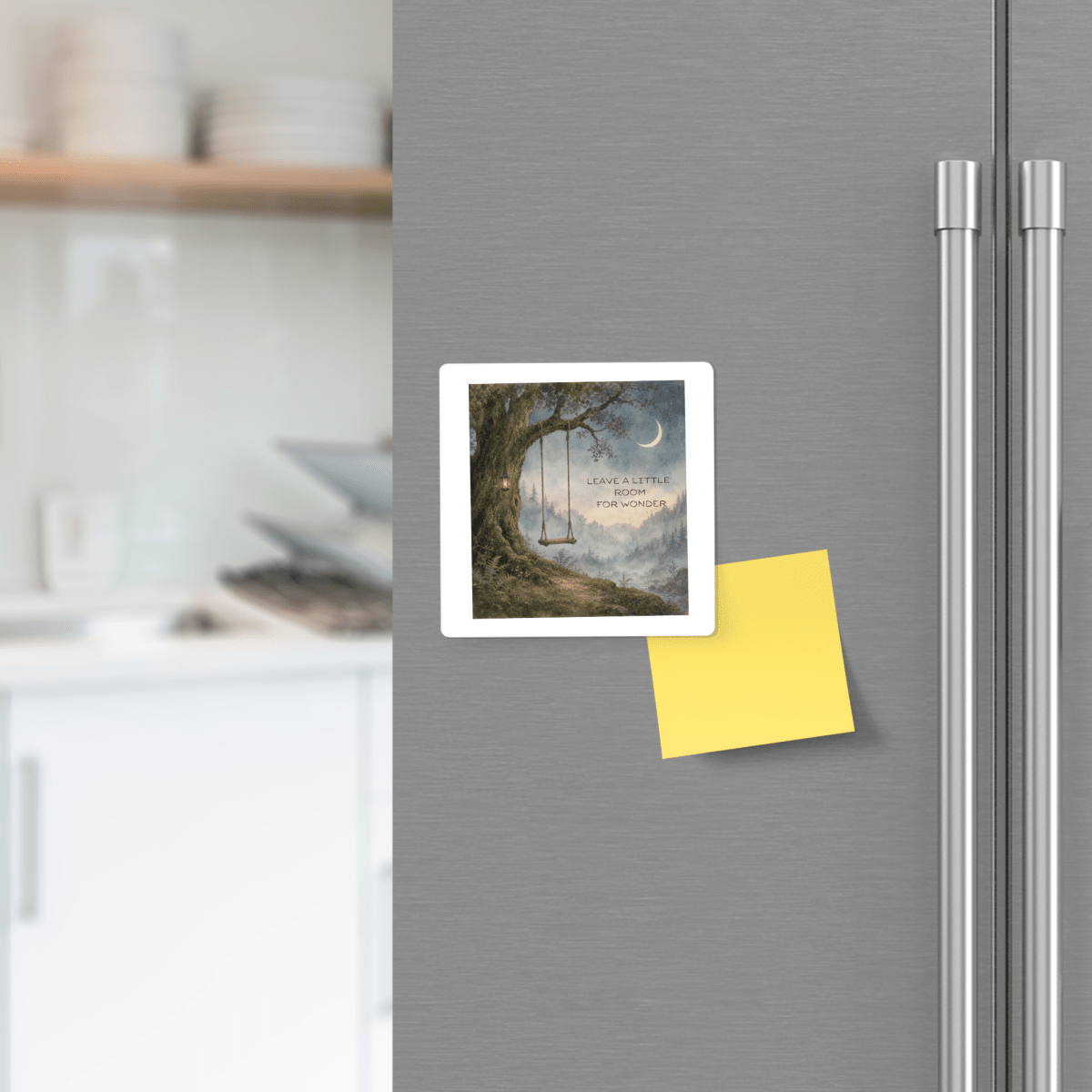 A fridge decorated with Mirabilia Boutique’s "Leave a Little Room for Wonder" magnet—designed to look like a yellow sticky note—and a square tree photograph. In the background, a modern kitchen with white dishes and countertops enhances the nature-inspired décor.