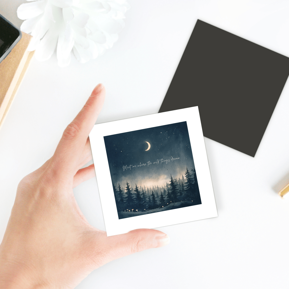 A hand holds the "Meet Me Where the Wild Things Dream Magnet" by Mirabilia Boutique, a square magnet depicting a moonlit night sky above a forest with cursive script—a perfect touch of celestial decor. Nearby on the white desk sits a black square and another nature-inspired magnet shaped like a white flower.