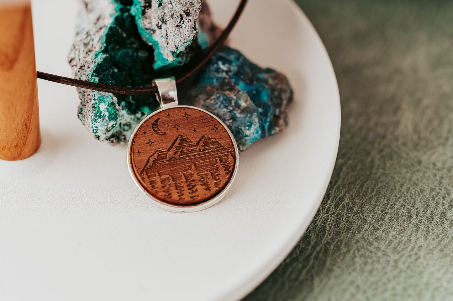 The Engraved Wood Pendant Necklace – Mountain Night Sky Design by Mirabilia Boutique features a round wood pendant with engraved mountains, trees, stars, and a crescent moon on a dark cord, shown on white with teal and blue crystals in the background.
